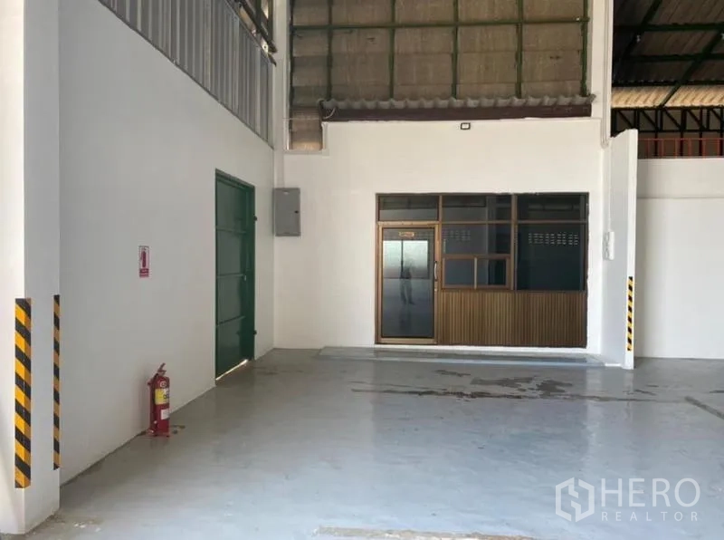 Warehouse for rent in Mueang Rayong, Rayong - Warehouse corner with a green service door, fire extinguisher, and glass-fronted office.