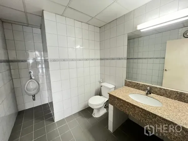Office Space for rent or sale - Clean tiled bathroom featuring a toilet, urinal, large mirror, and granite countertop sink.