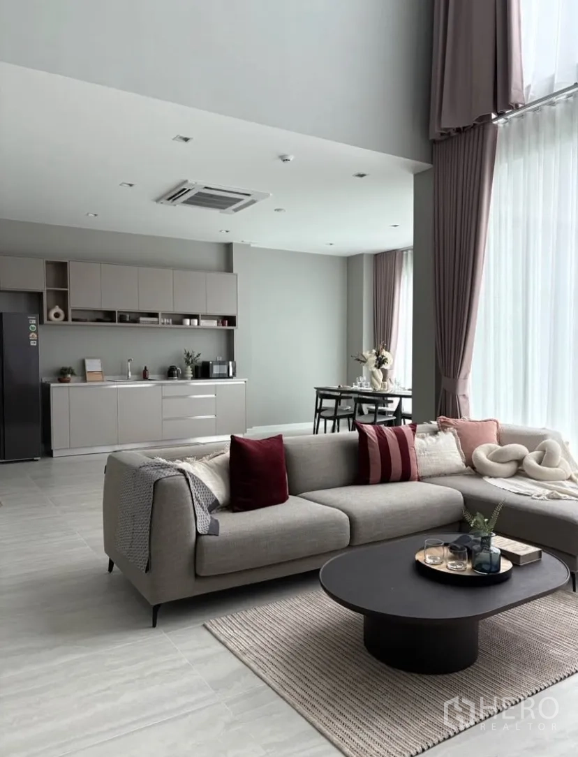 Detached House for rent in Suan Luang, Bangkok - Bright double-height living room with sectional sofa, floor-to-ceiling curtains and a minimalist pantry kitchen.