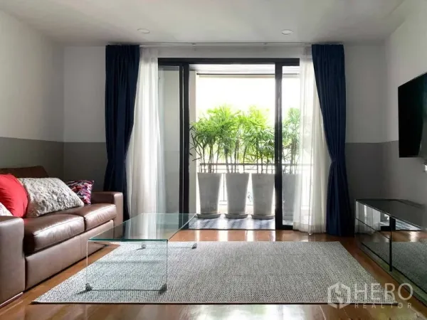 Condominium for sale in Watthana, Bangkok - Living room opening to a small balcony with potted plants, floor-to-ceiling curtains and wall-mounted TV.