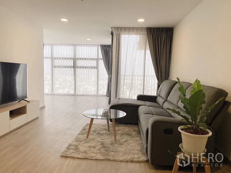 คอนโด ให้เช่า - ห้องนั่งเล่นสว่าง โซฟา L-Shape ทีวี และกระจกเต็มบานมุมห้องมองเห็นวิวเมือง