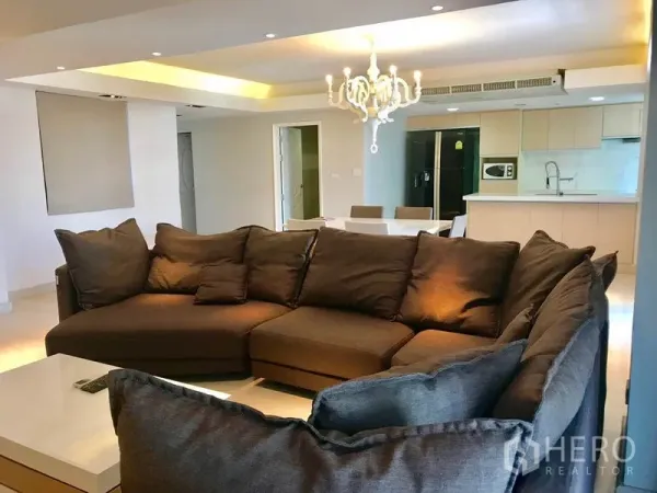 Condominium for rent or sale in Watthana, Bangkok - Large L-shaped sofa in an open-plan living room with chandelier, dining table, and kitchen behind.