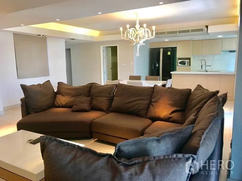 Condominium for rent or sale in Watthana, Bangkok - Large L-shaped sofa in an open-plan living room with chandelier, dining table, and kitchen behind.