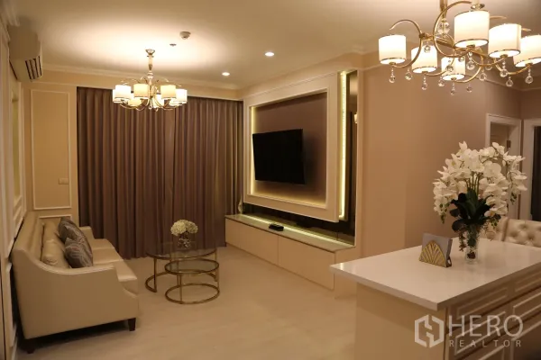 Condominium for rent in Huai Khwang, Bangkok - Warm-toned living room with beige sofa, chandelier lighting, framed TV feature wall, glass nesting coffee tables, full-length curtains and a counter with flowers.