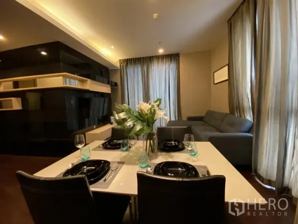 Condominium for rent in Watthana, Bangkok - Open-plan living and dining area with set table, L-shaped sofa, built-in shelves and floor-to-ceiling curtains.