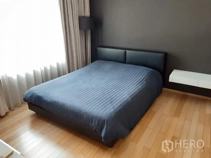 Condominium for rent in Khlong Toei, Bangkok - Spacious bedroom featuring a large bed, dark feature wall, wood floors and sheer curtains by the window.