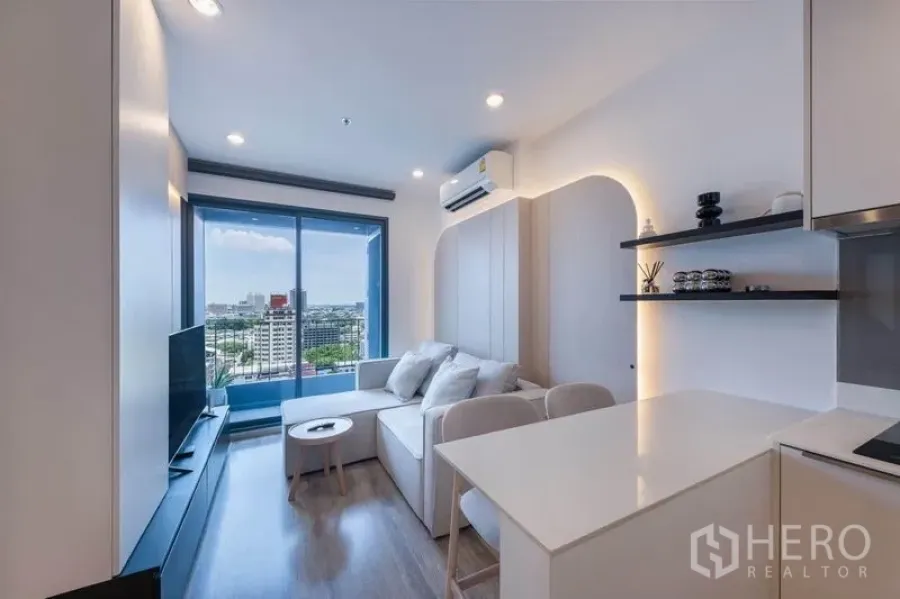 Condominium for rent in Bang Na, Bangkok - Bright living room with L-shaped sofa, TV cabinet, and floor-to-ceiling sliding doors opening to a balcony with city view.