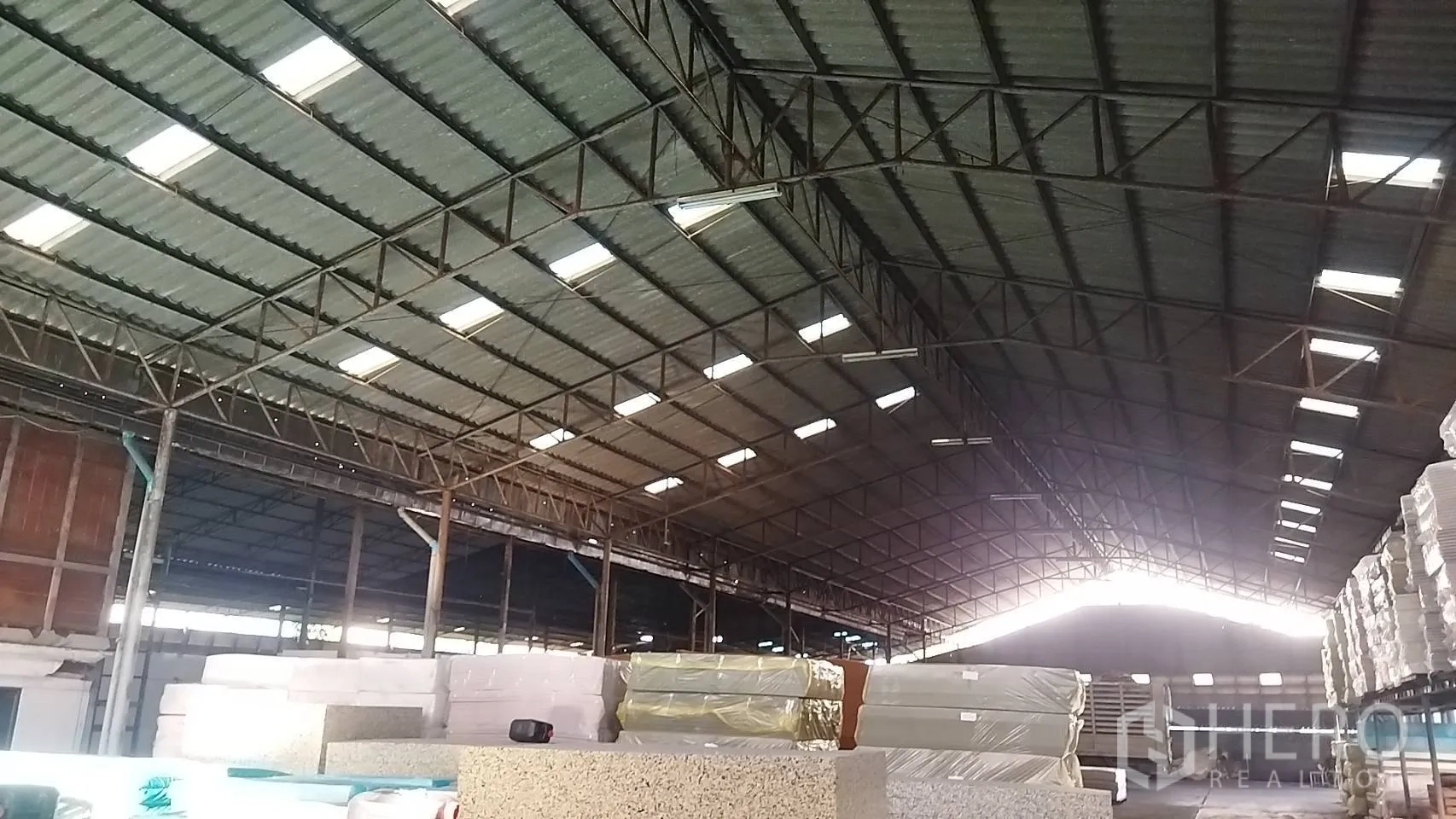 Warehouse for rent in Bang Khun Thian, Bangkok - View toward the ridgeline roof with trusses and inventory stacks under bright skylights.