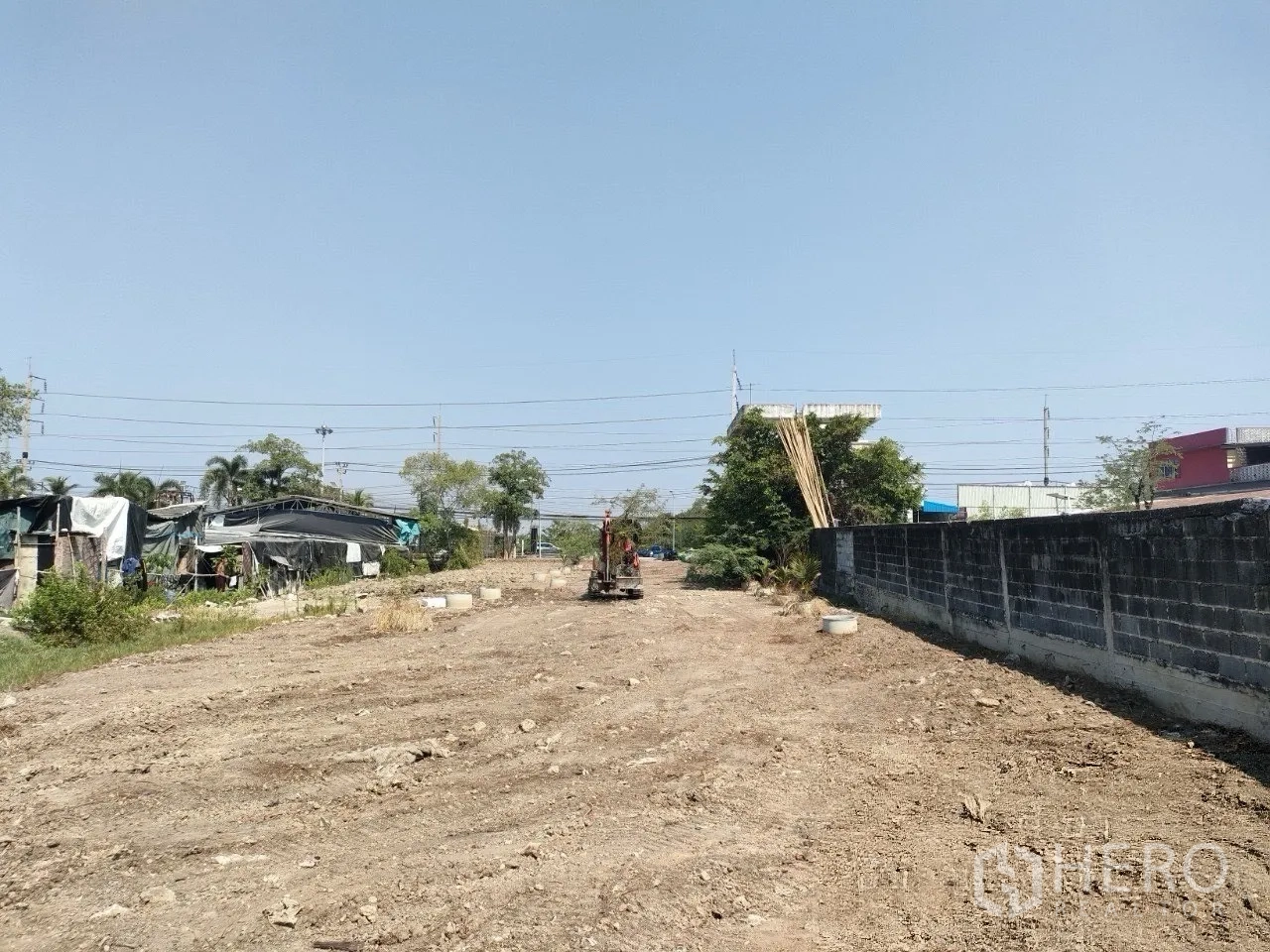 Land for sale in Mueang Samut Prakan, Samut Prakan - Dirt surface land beside a masonry wall with utility lines and makeshift sheds in the background.