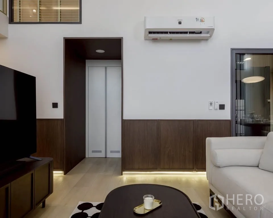 Condominium for rent in Khlong Toei, Bangkok - Living area with sofa, TV console, wood paneling and a wall-mounted air conditioner.