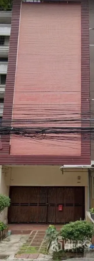 อาคารพาณิชย์ ให้เช่าในKhlong Toei, Bangkok - ด้านหน้าอาคารพาณิชย์สูงสี่ชั้น มีประตูม้วนสีชมพูเต็มบาน ประตูไม้ด้านล่าง สายไฟพาดเหนือถนน และต้นไม้เล็กหน้าบ้าน