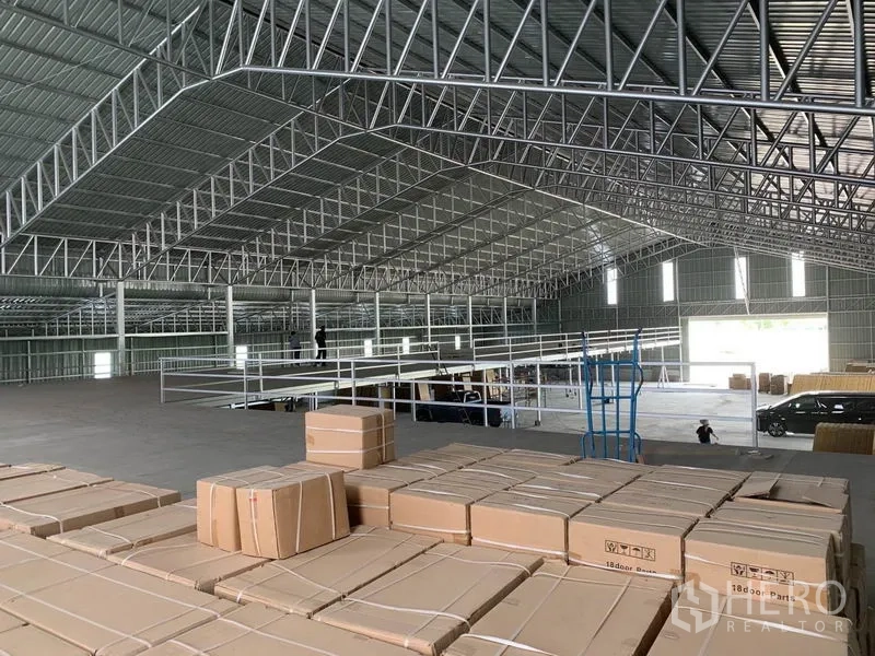 Warehouse for sale in Phra Nakhon Si Ayutthaya - High-clearance warehouse interior with steel truss roof and mezzanine, stacked with boxed inventory.