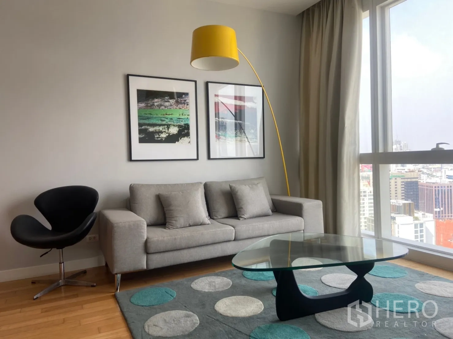 คอนโด ให้เช่าหรือขายในKhlong Toei, Bangkok - มุมพักผ่อนพร้อมโซฟาสีเทา งานศิลป์ พรมลายจุด และหน้าต่างบานใหญ่เห็นวิวกรุงเทพฯ