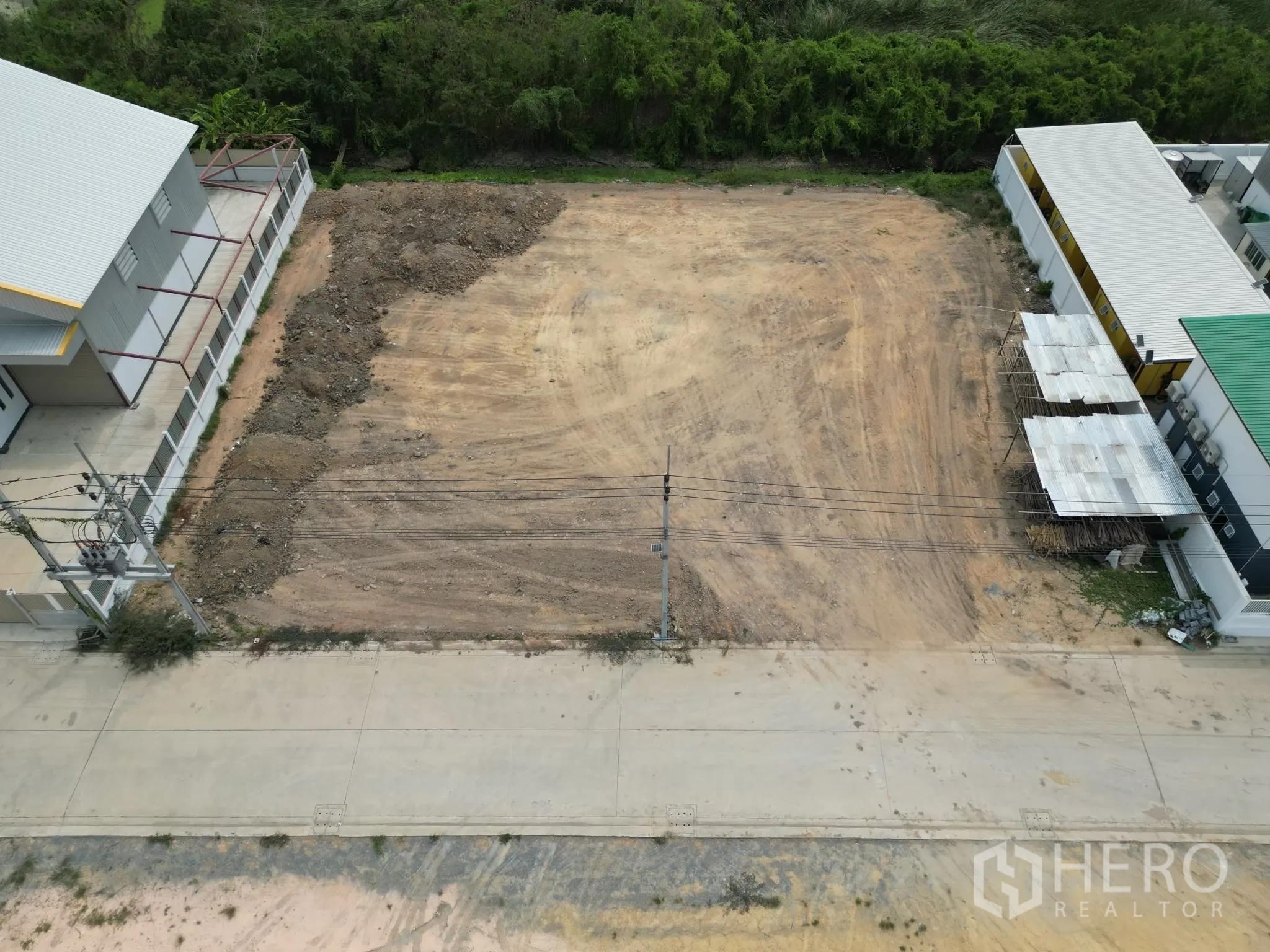 Land for sale in Sai Noi, Nonthaburi - Aerial view of a cleared, leveled land plot bordered by warehouses and a concrete access road with power lines.