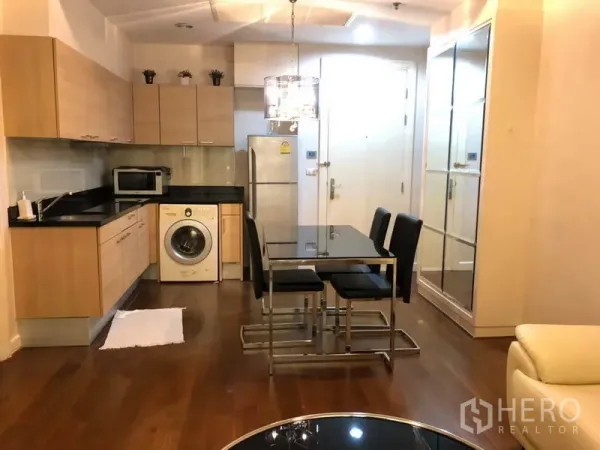 公寓 出租在Pathum Wan, Bangkok - 开放式厨房，浅色橱柜与黑色台面，配备家电、洗衣机及吊灯下的玻璃餐桌。