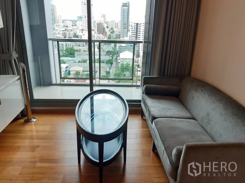 Condominium for rent in Watthana, Bangkok - Living corner with gray sofa and round glass coffee table facing a city-view balcony.