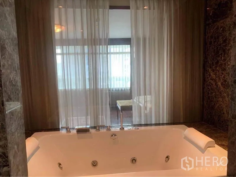 Condominium for rent in Watthana, Bangkok - Marble bathroom featuring a large jacuzzi bathtub with headrests and a glass partition with sheer curtains.