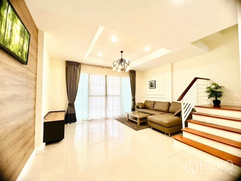 联排别墅 出租在Prawet, Bangkok - 采光充足的客厅，高挑天花、落地窗与木质装饰墙