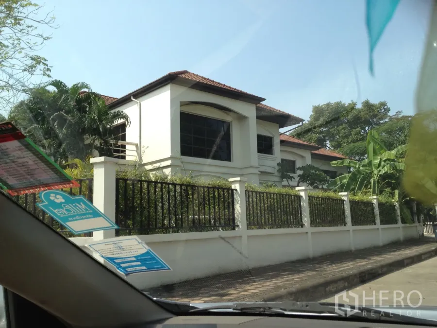 独立式住宅 出租在Nonthaburi - 两层白色独栋别墅，棕色瓦顶，大面积窗户与黑色金属围栏，绿树环绕，位于暖武里帕克烈。
