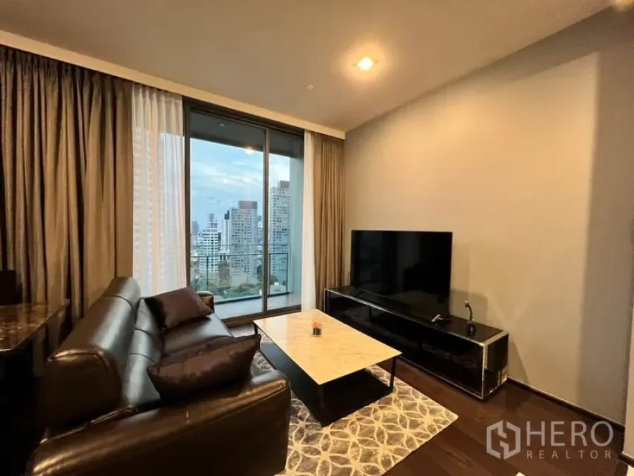 Condominium for rent in Watthana, Bangkok - Living room with leather sofa, marble coffee table, large TV and floor-to-ceiling window opening to a city-view balcony.