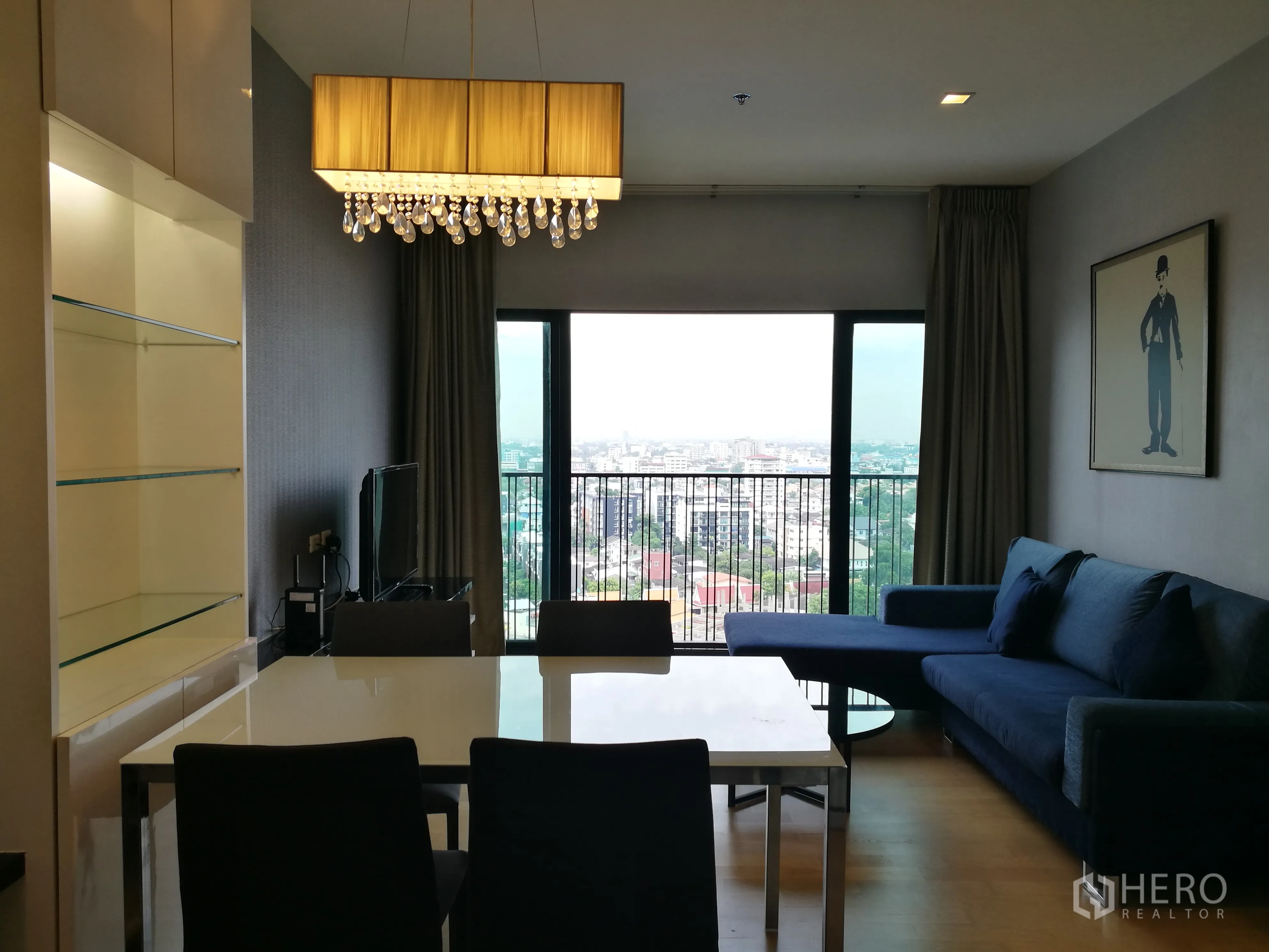 Condominium for rent in Watthana, Bangkok - Open‑plan living and dining area with crystal chandelier, glass display shelves, dining set, and balcony doors framing a city view.
