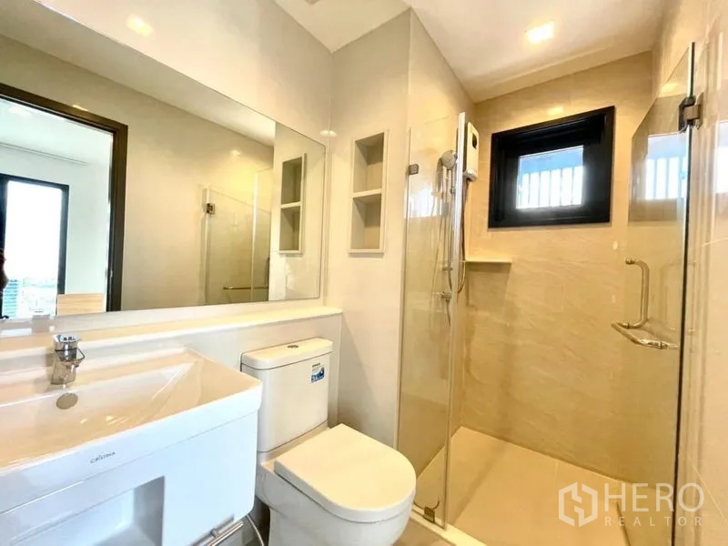 Condominium for rent in Chatuchak, Bangkok - Modern bathroom with large mirror, toilet and glass-enclosed shower with window.