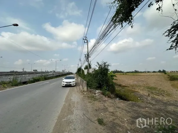 Land for sale in Bang Bua Thong, Nonthaburi - Two‑lane road with a parked car, overhead cables and the open land parcel stretching to the right.