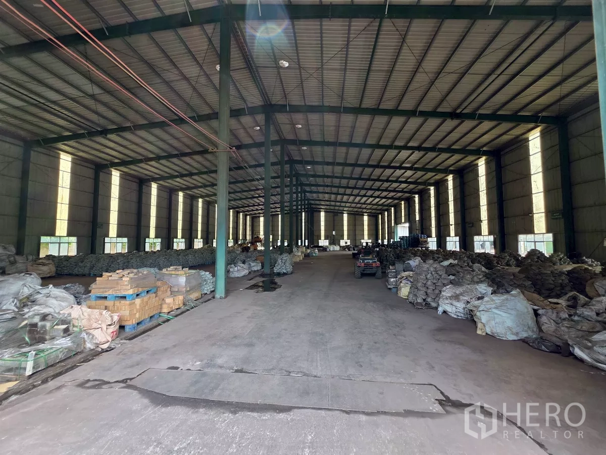 Factory for rent in Mueang Samut Sakhon, Samut Sakhon - High‑clear factory hall with steel columns, roof lighting and organized bulk storage on a concrete floor.