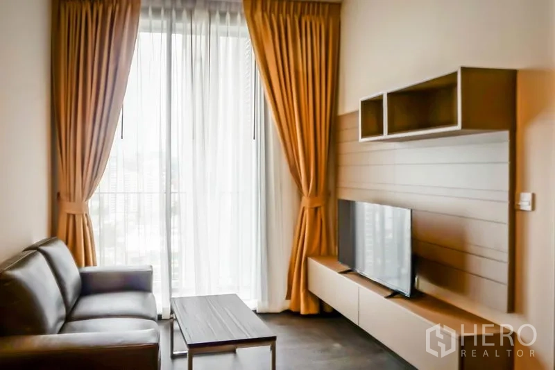 Condominium for sale in Watthana, Bangkok - Bright living room with leather sofa, TV console and floor-to-ceiling window with curtains.