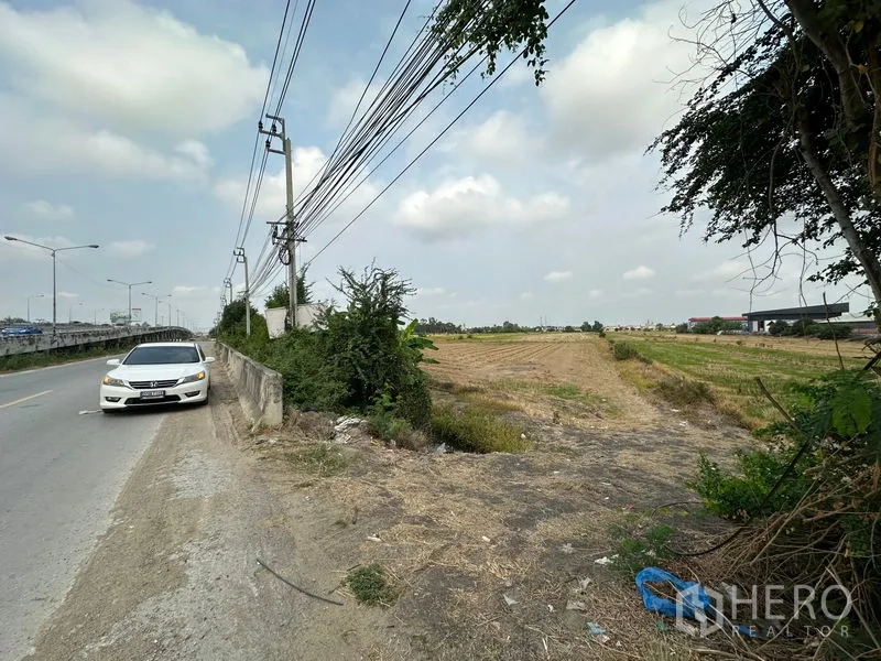 Land for sale in Bang Bua Thong, Nonthaburi - Entrance to a large flat field beside the paved road with utility poles and drainage, suitable for development.