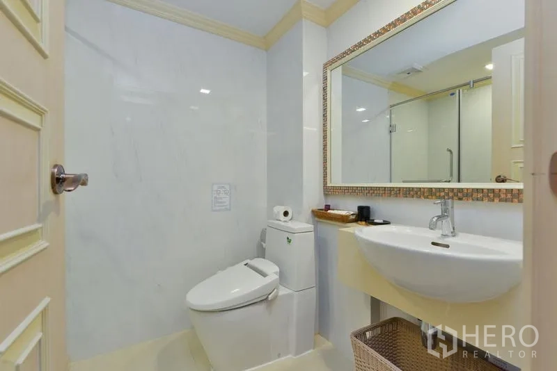Apartment for rent in Khlong Toei, Bangkok - Bright bathroom with large mirror, vanity sink, toilet and glass shower enclosure.