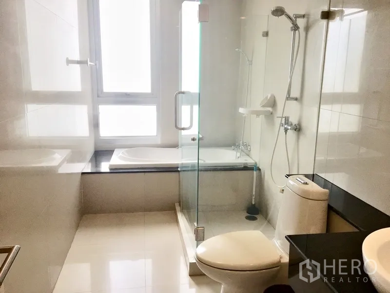 Condominium for rent in Khlong Toei, Bangkok - Modern bathroom featuring a bathtub by the window and a glass-enclosed shower.