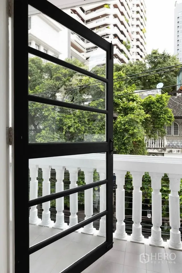 联排别墅 出租在Khlong Toei, Bangkok - 小阳台配白色栏杆与黑框门，可眺望树木与周边建筑。