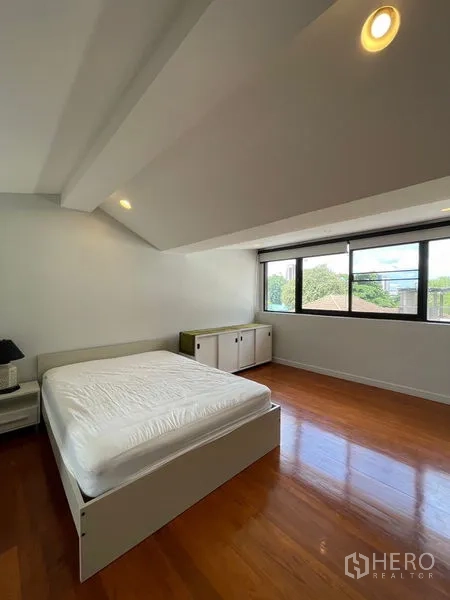 Townhouse for rent in Khlong Toei, Bangkok - Bedroom with sloped ceiling, wide window and polished wood floors.