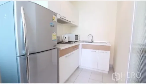 Condominium for rent in Watthana, Bangkok - Compact L-shaped kitchen with white cabinets, stainless refrigerator, microwave and sink.