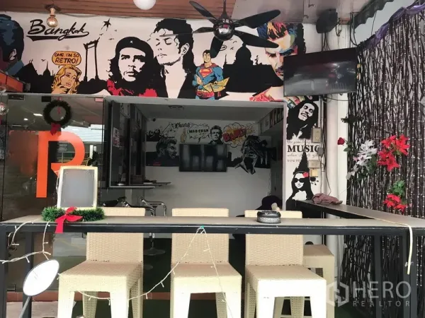 零售空间 出租在Watthana, Bangkok - 开放式铺面，藤编高脚椅、吊扇、壁挂电视与装饰藤条，背景为流行艺术壁画。
