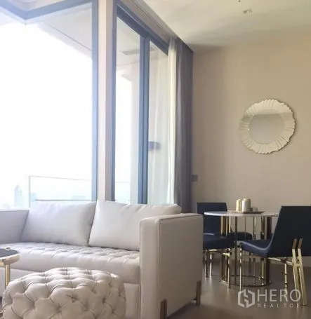Condominium for rent in Watthana, Bangkok - Bright living and dining area with white sofa, round mirror and floor‑to‑ceiling windows opening to a balcony.