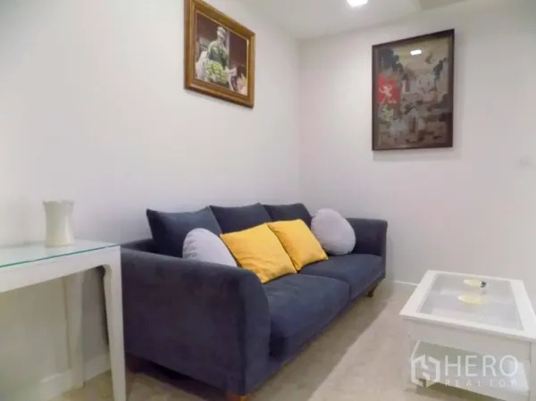 Condominium for rent or sale in Khlong Toei, Bangkok - Cozy living area with dark fabric sofa, colorful cushions, wall art and white coffee table under recessed lights.