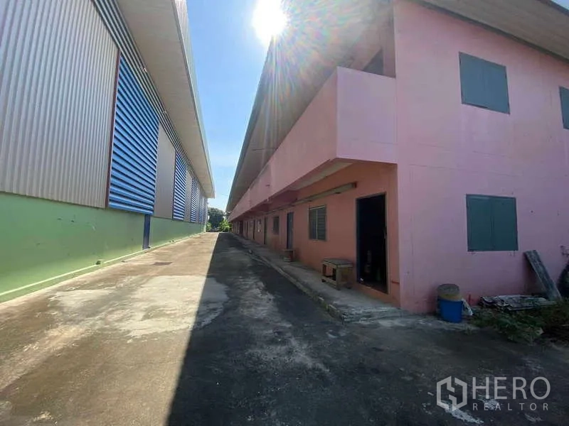 Warehouse for rent in Lat Lum Kaeo, Pathum Thani - Narrow service lane between warehouse and pink staff quarters with sunlight overhead.