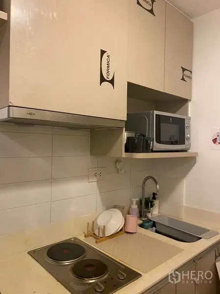 Condominium for sale in Bang Rak, Bangkok - Compact kitchenette with electric cooktop, sink, microwave and overhead cabinets in neutral tones.