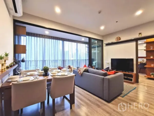 Condominium for rent in Huai Khwang, Bangkok - Open‑plan living and dining area with grey sofa, set dining table, floor‑to‑ceiling windows and a balcony city view in a Bangkok condo.