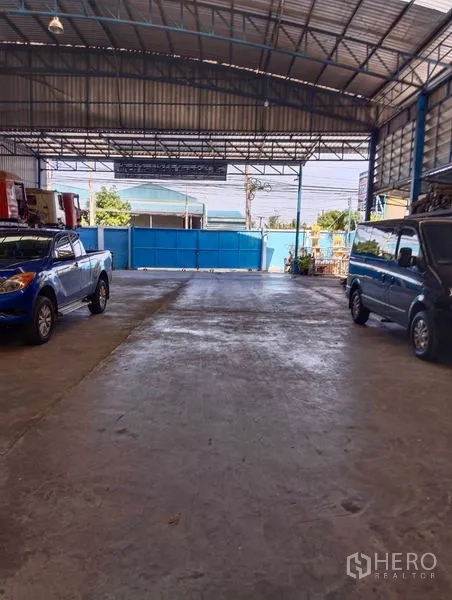 Warehouse for sale in Sai Noi, Nonthaburi - Drive-in warehouse entrance with blue gate, concrete apron, and parked vehicles under roof.