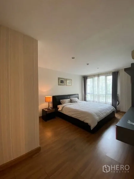 Condominium for rent in Watthana, Bangkok - Spacious bedroom with large window, curtains, bedside lamp and wood flooring.