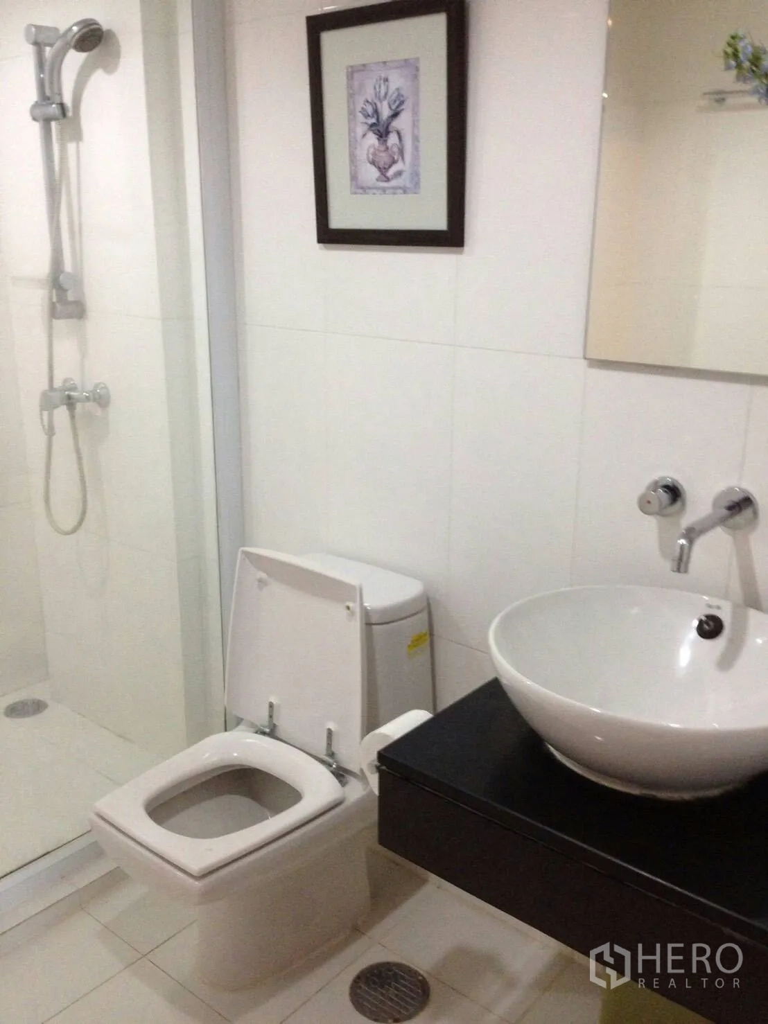 Condominium for rent in Pathum Wan, Bangkok - Modern white-tiled bathroom with glass shower, toilet and round vessel sink.