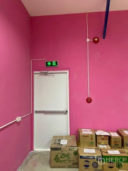 Warehouse for rent in Khlong Luang, Pathum Thani - Fire exit door with emergency sign and alarm on pink wall, boxes stacked nearby.