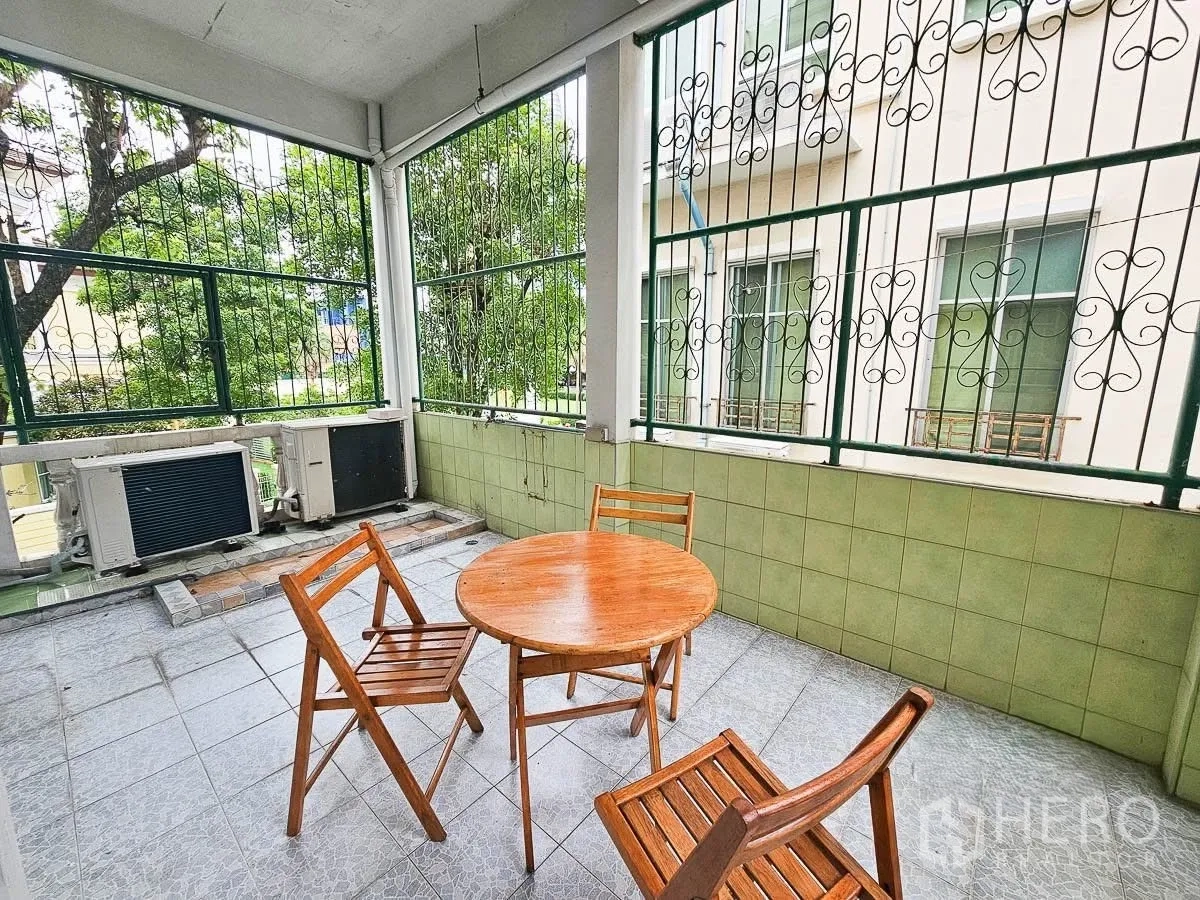联排别墅 for rent in Yan Nawa, Bangkok - 覆盖式阳台，木质圆桌配三把椅子，绿树景观与空调室外机。