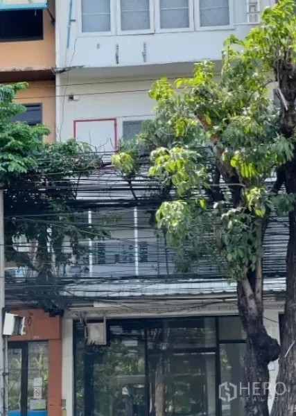 店屋 出售在Watthana, Bangkok - 瓦塔那区素坤逸71上的三层店屋外立面，宽幅玻璃门面、金属雨棚、阳台窗及街道行道树与电缆。