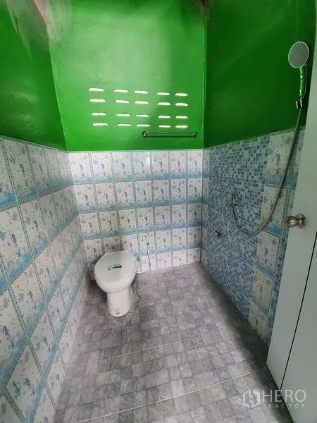 Warehouse for rent in Nonthaburi - Tiled bathroom with toilet and handheld shower under a green ceiling with ventilation blocks.