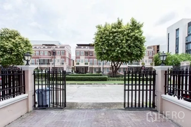 Townhouse for rent in Watthana, Bangkok - View from the driveway through black iron gate to a tree-lined street and facing townhouses.