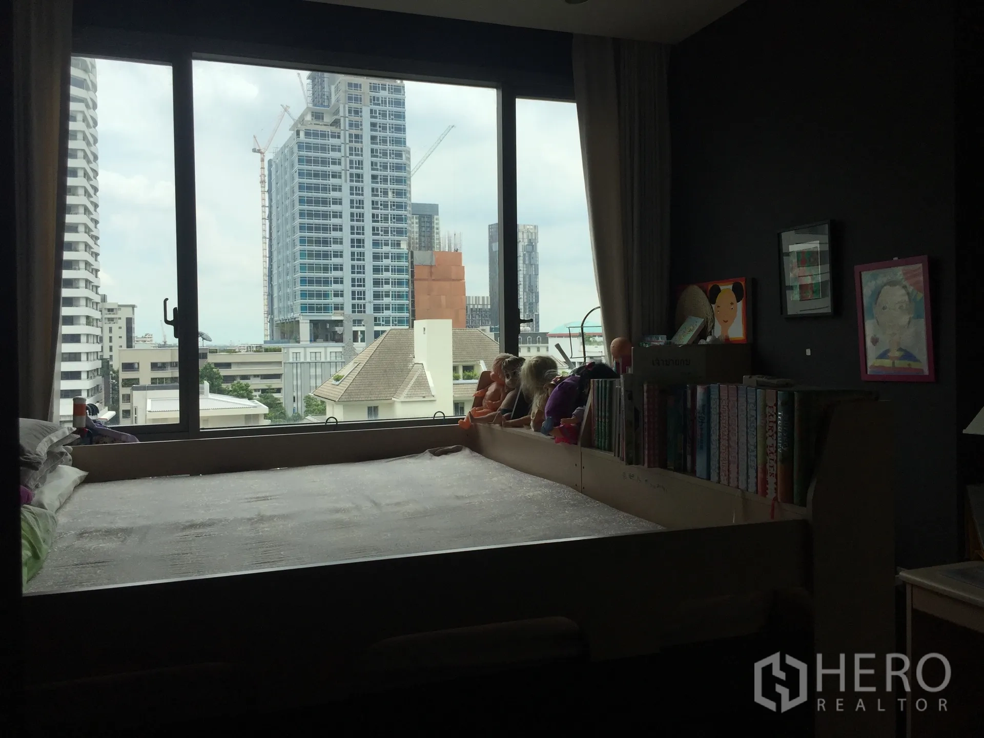Condominium for sale in Watthana, Bangkok - Bedroom with large window, platform bed, bookshelf, and skyline view of high-rise buildings.